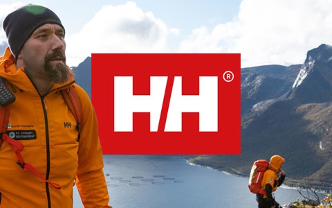 A man wearing a orange Helly Hansen jacket, a scenery of mountains touching the clouds and the Helly Hansen logo in the centre
